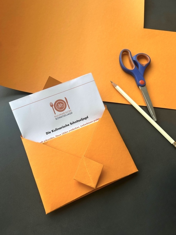 Ein selbstgebastelter Origami-Umschlag in orange. Ein Gutschein für die Kulinarische Schnitzeljagd ragt aus dem Umschlag hervor.
