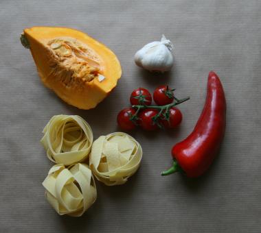 Die einzelnen Zutaten Nudeln, Tomaten, Paprika, Kürbis und Knoblauch auf grauem Untergrund