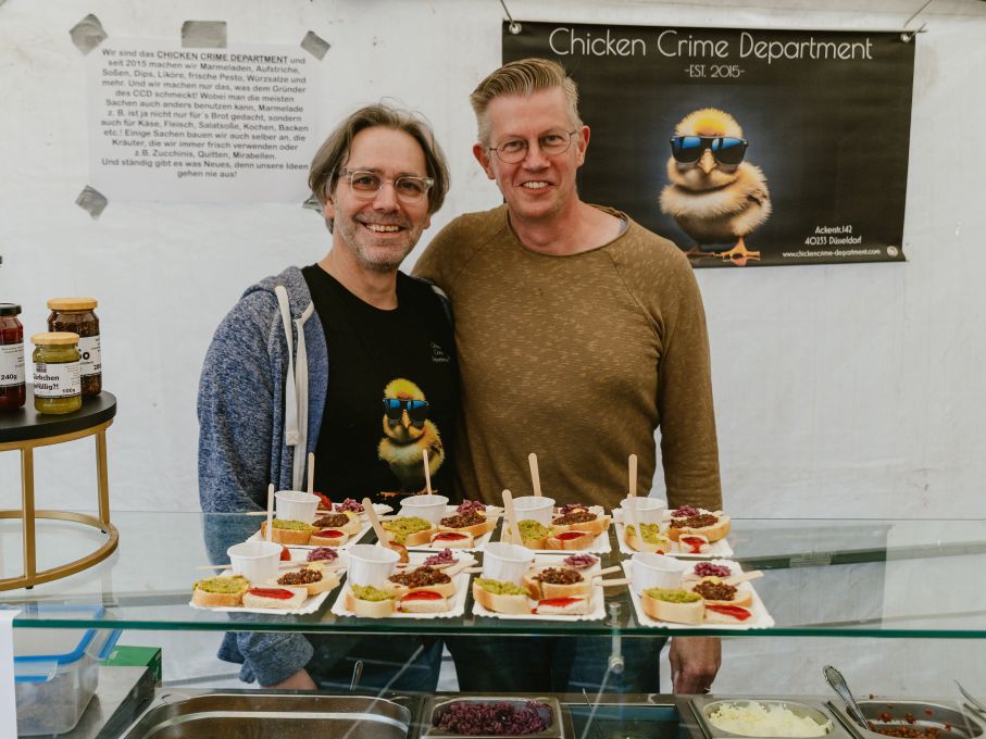 Die Inhaber des Feinkostgeschäfts Chicken Crime Department zusammen hinter dem Tresen ihres aufgebauten Genusszeltes für die Kulinarische Schnitzeljagd. Auf dem Tresen befinden sich die vorbereiteten Kostproben.