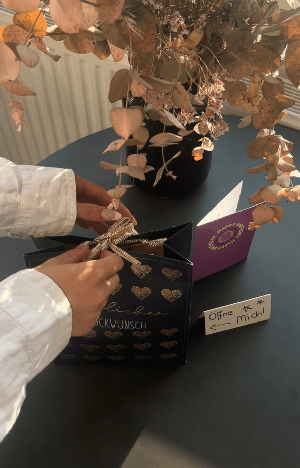 Hände, die eine Wundertüte öffnen. Im Hintergrund sind Blumen, eine Geburtstagskarte und ein Schild mit dem Aufzug "Öffne mich" zu sehen.
